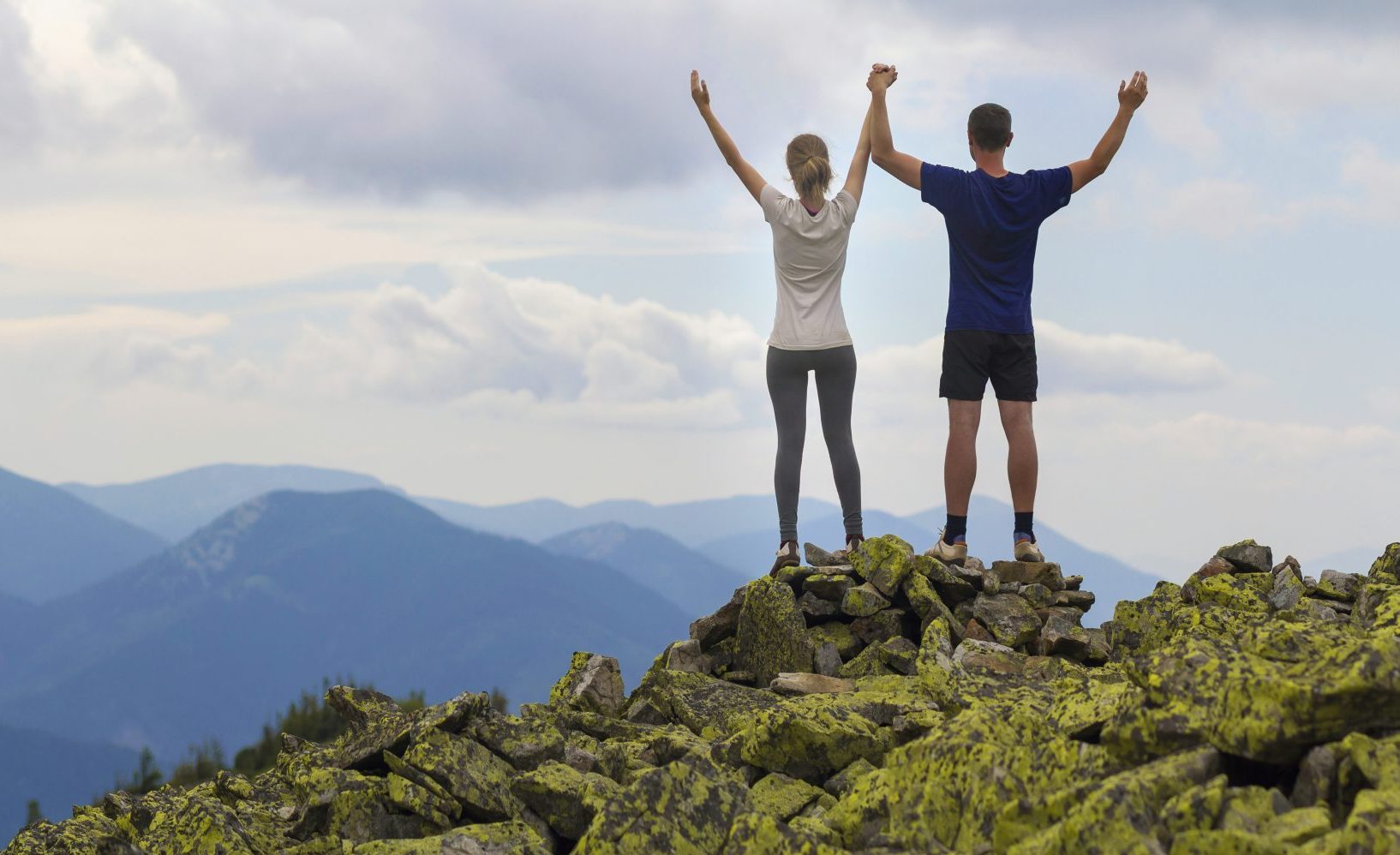 Back view of young couple, athletic boy and slim girl standing with raised arms on rocky mountain top enjoying breathtaking summer mountain view. Tourism, traveling and healthy lifestyle concept.